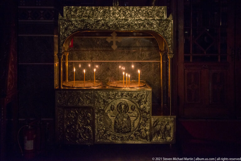 Burning votive candles on metal trays beneath an ornate dark canopy with a cross.