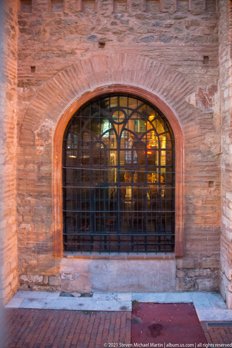 Large arched window with black metal grille set into thick weathered brick masonry.