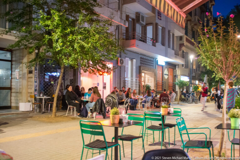 Green café tables and chairs on a busy pedestrian street with diners and illuminated storefronts at night.