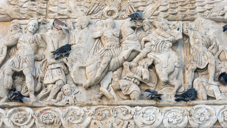 Crowded Roman battle relief with mounted soldiers, fallen figures, and pigeons perched on the carved marble.