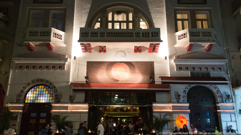 Illuminated façade with balconies, stained-glass arches, striped awnings, and people entering at night.