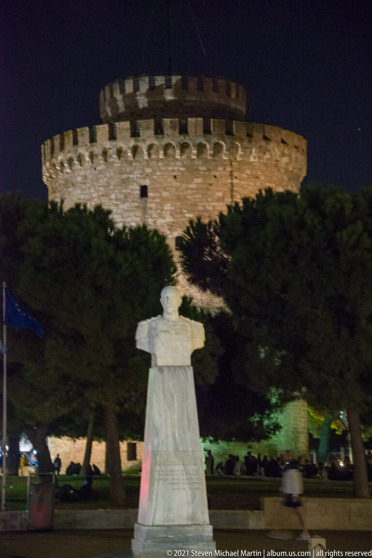 Illuminated White Tower behind trees and a pale stone monument at night.