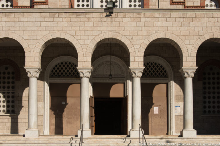 Five-arched church portico with pale columns, central dark doorway, steps, and patterned window screens.