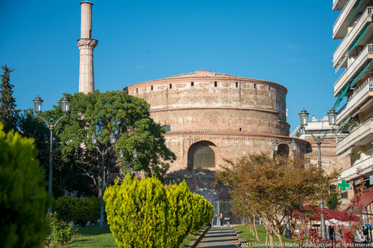 Cylindrical brick Rotunda of Galerius beside its tall surviving minaret, seen beyond trees and a walkway.
