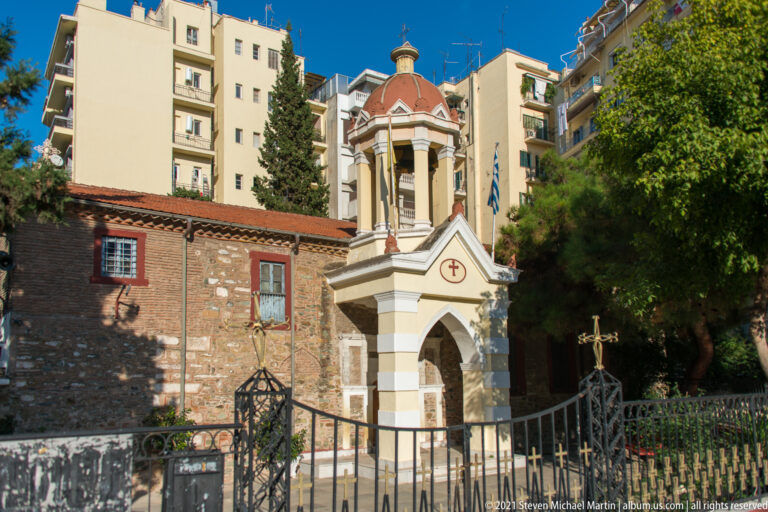 Small stone-and-brick Orthodox church with a domed porch, crosses, trees, and apartment buildings behind it.