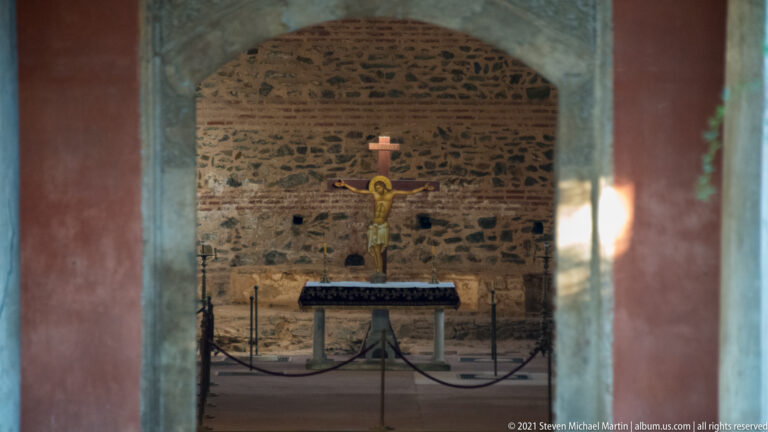 Crucifix and simple altar centered in a rough stone-walled sanctuary viewed through an opening.