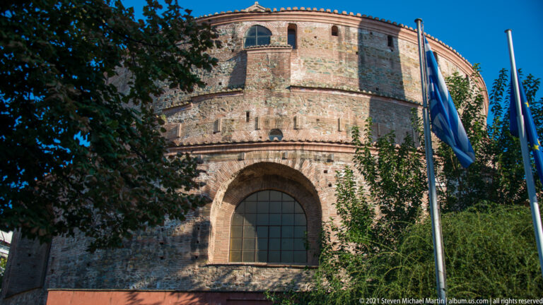 Curved brick exterior of the Rotunda with a large arched window, trees, and Greek and EU flags.