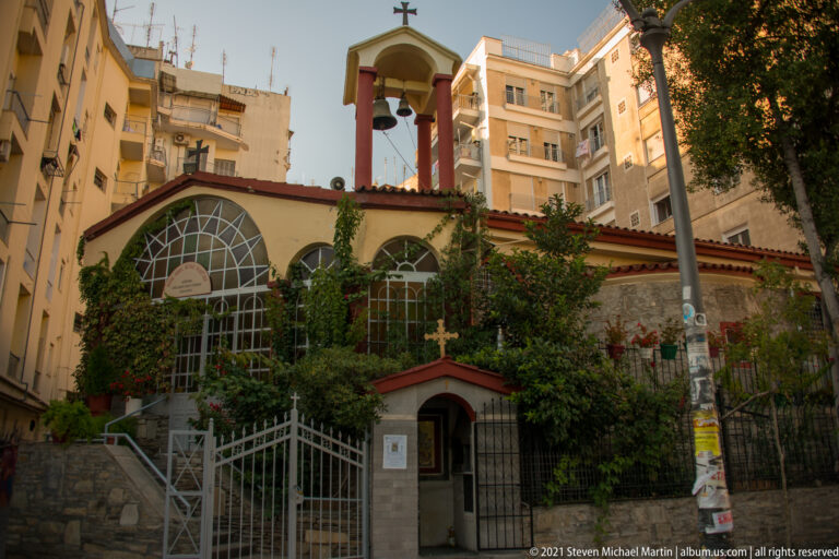 Small Orthodox chapel with arched windows, crosses, vines, and a bell frame among apartment buildings.