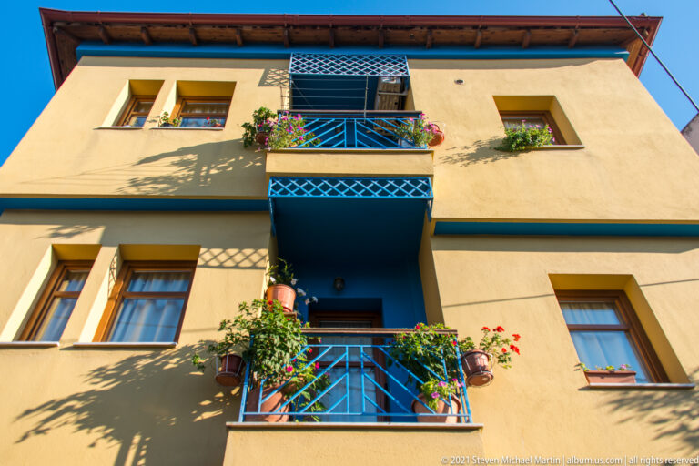 Blue metal balconies filled with flowering plants on a sunlit ochre residential façade.