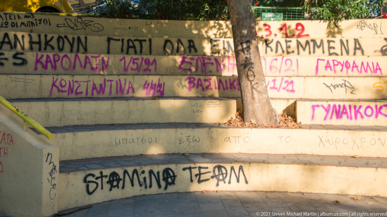 Concrete steps covered with handwritten Greek text, dates, names, and black and magenta graffiti beneath a tree.