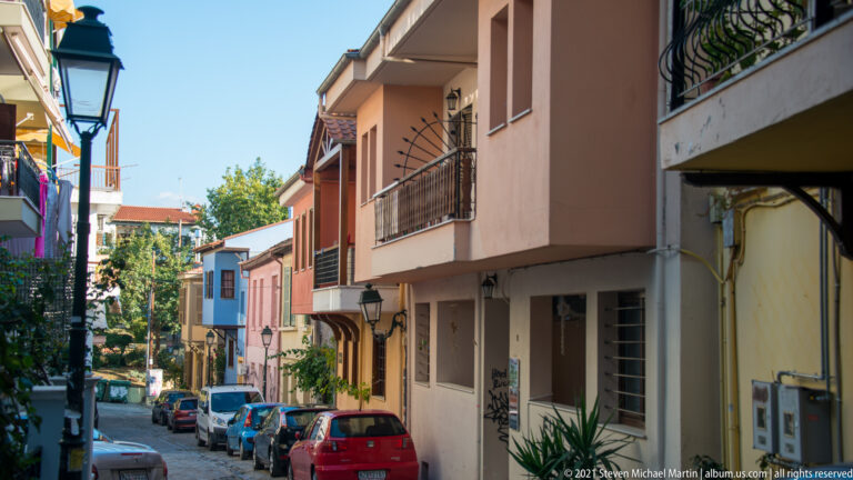 Narrow residential street with parked cars, balconies, stucco houses, trees, and tiled roofs in Thessaloniki.