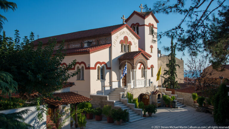 Small white Orthodox church with red trim, arched entrance, crosses, steps, and potted plants.