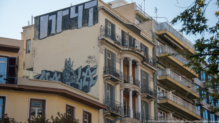 Tall ornamented residential building with balconies above a lower corner façade covered in graffiti.