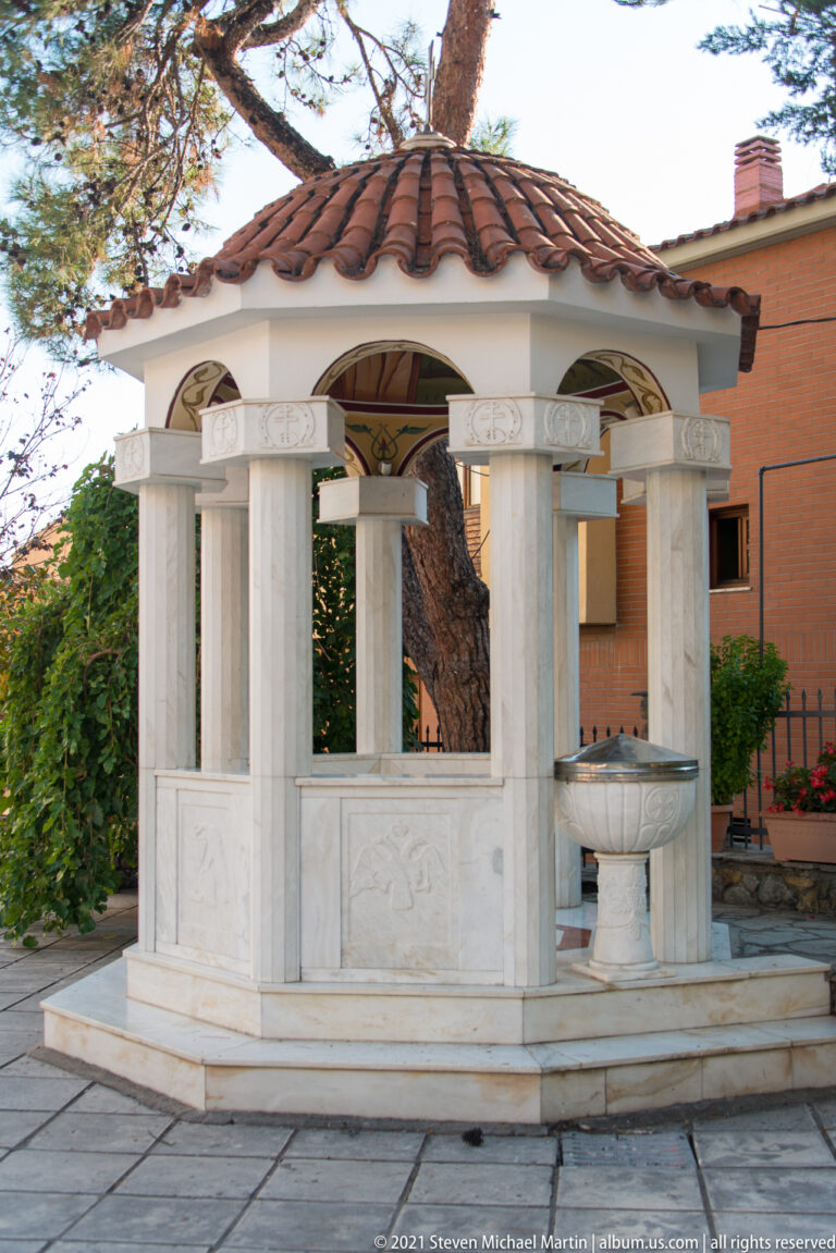 Open marble fountain pavilion with slender columns, carved capitals, red-tiled roof, and basin at right.