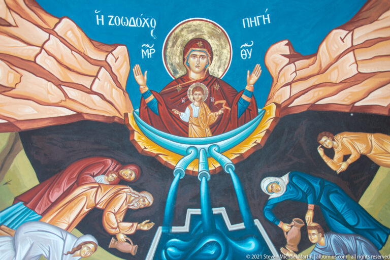 Colorful Orthodox mural with Virgin and Child above flowing water, surrounded by figures in a rocky landscape.
