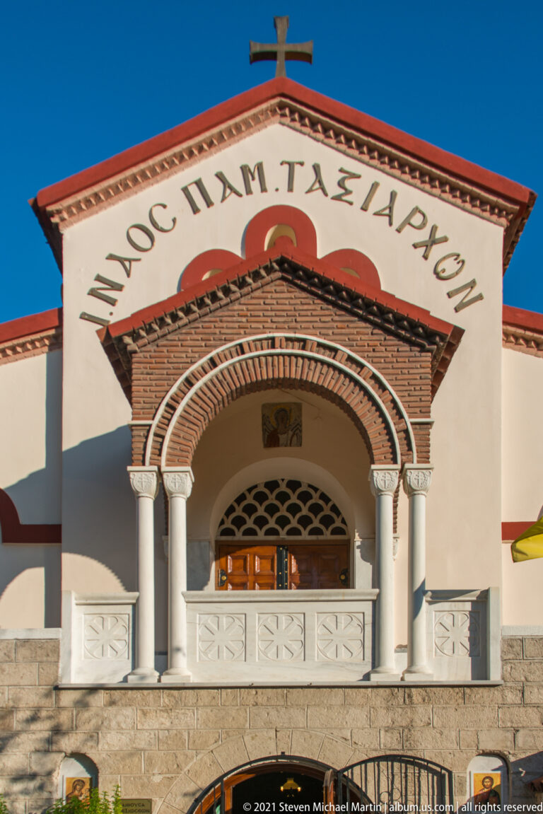 White Orthodox church façade with brick arched entrance, paired columns, Greek dedication, tiled roof, and cross.