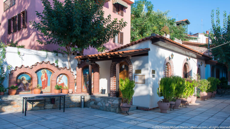 Small white chapel with red-tile roof, brick-trimmed windows, potted plants, and religious paintings in a paved courtyard.