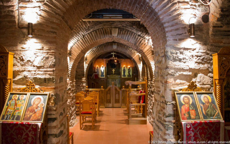Narrow stone-vaulted Orthodox interior with repeated arches, icons on lecterns, chairs, and a wooden iconostasis.