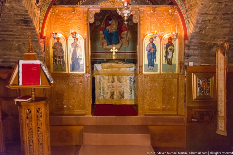 Carved wooden iconostasis with painted icons, central altar opening, red lectern, and stone wall behind.