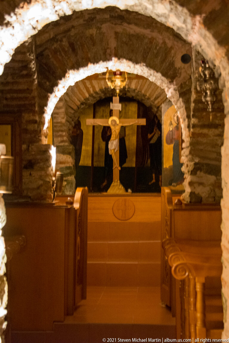 Crucifix beyond a low stone-and-brick arch with wooden benches lining the small church interior.