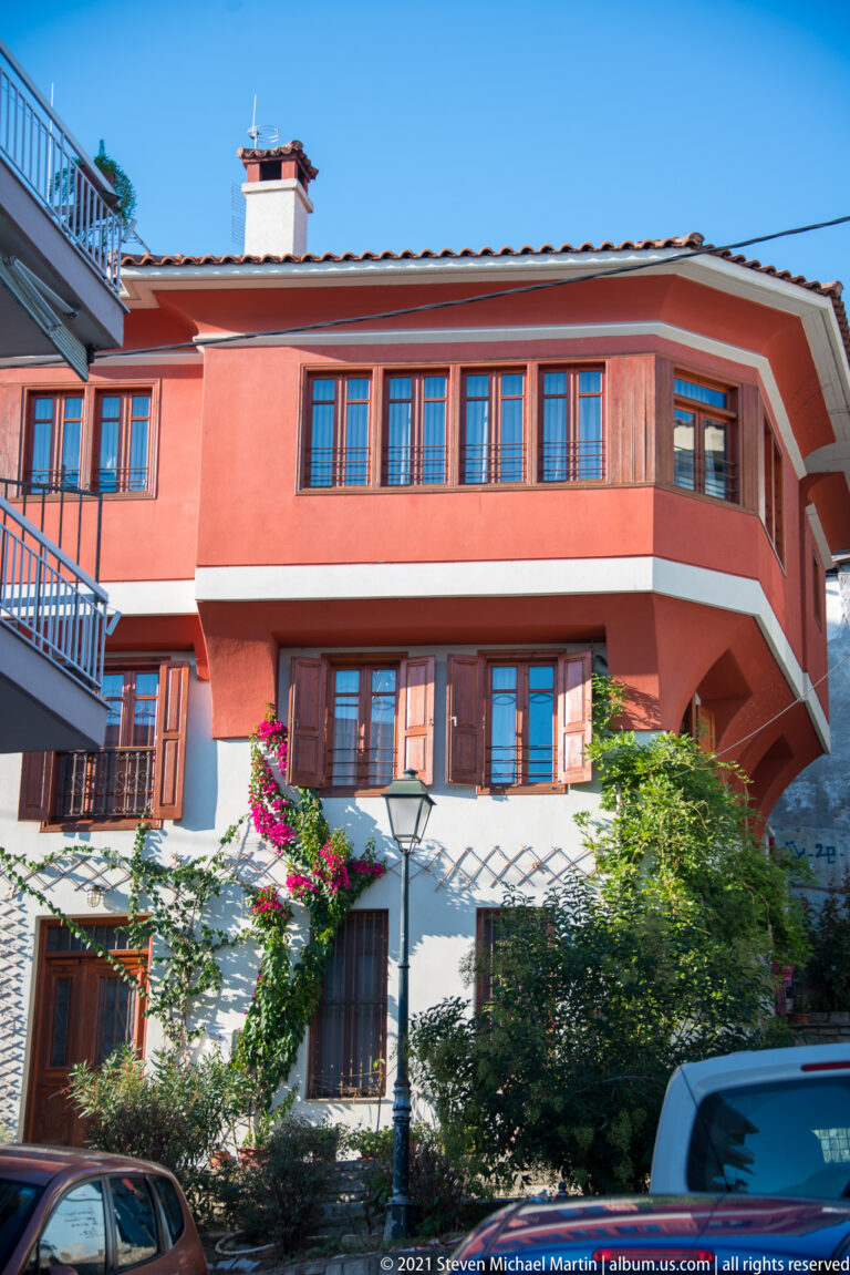 Coral-colored hillside residence with balconies, shutters, greenery, and parked car on a narrow street.
