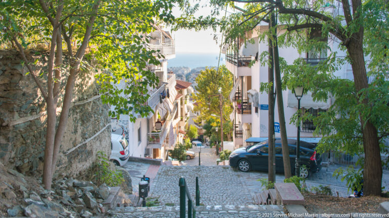 Steep cobbled residential lane descending between trees and houses toward a distant view of the sea.