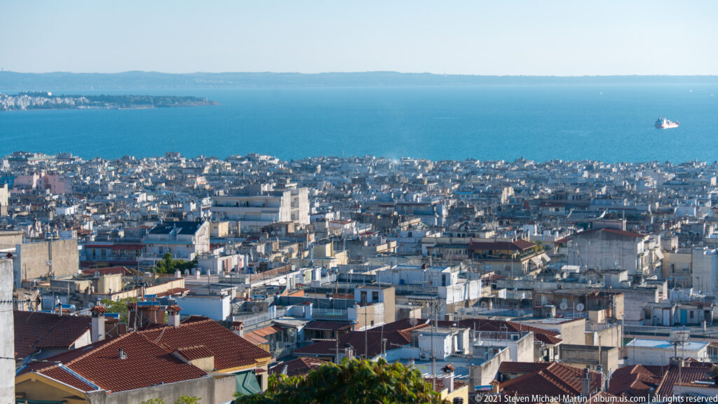 Elevated panorama over dense Thessaloniki rooftops to the blue Thermaic Gulf with a ship offshore.