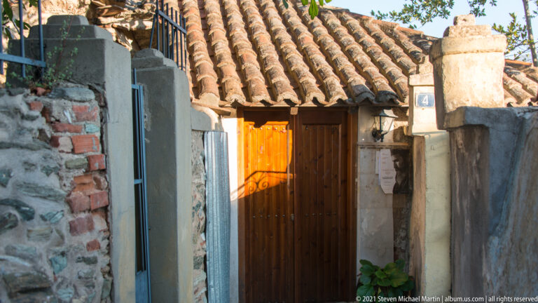 Wooden doorway beneath old clay roof tiles, surrounded by weathered brick, stone, and plaster walls.