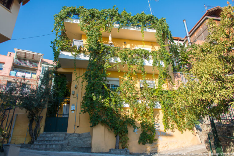 Yellow hillside house covered with vines and trees, with exterior steps and apartment buildings behind.