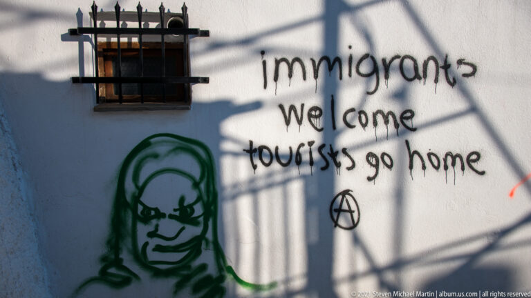 Graffiti on white wall reading “immigrants welcome tourists go home,” with anarchist symbol and green face stencil.