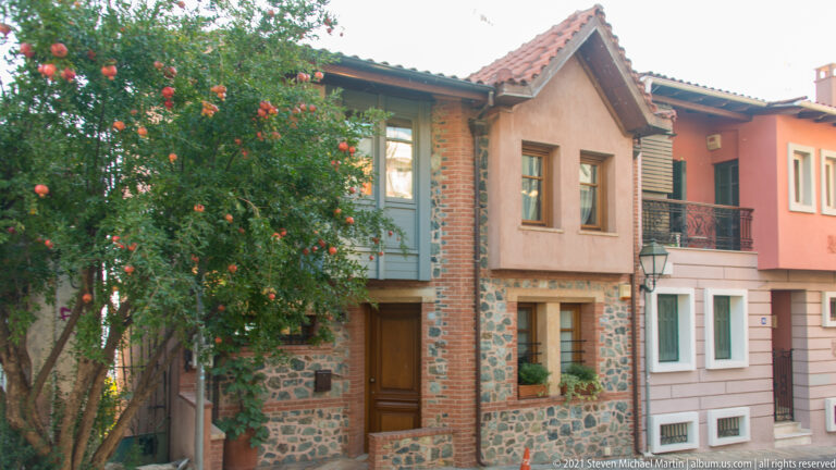 Traditional two-story house with stone-and-brick masonry, timber trim, tiled roof, shutters, and flowering tree.