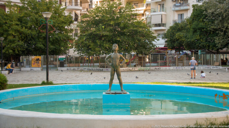 Standing female statue centered in a shallow circular fountain in a tree-lined urban square.
