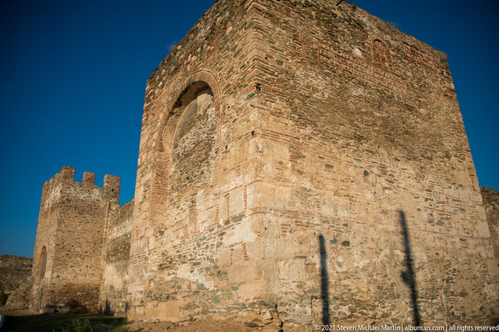 Massive rectangular stone-and-brick fortification tower with arched opening and crenellated wall behind.