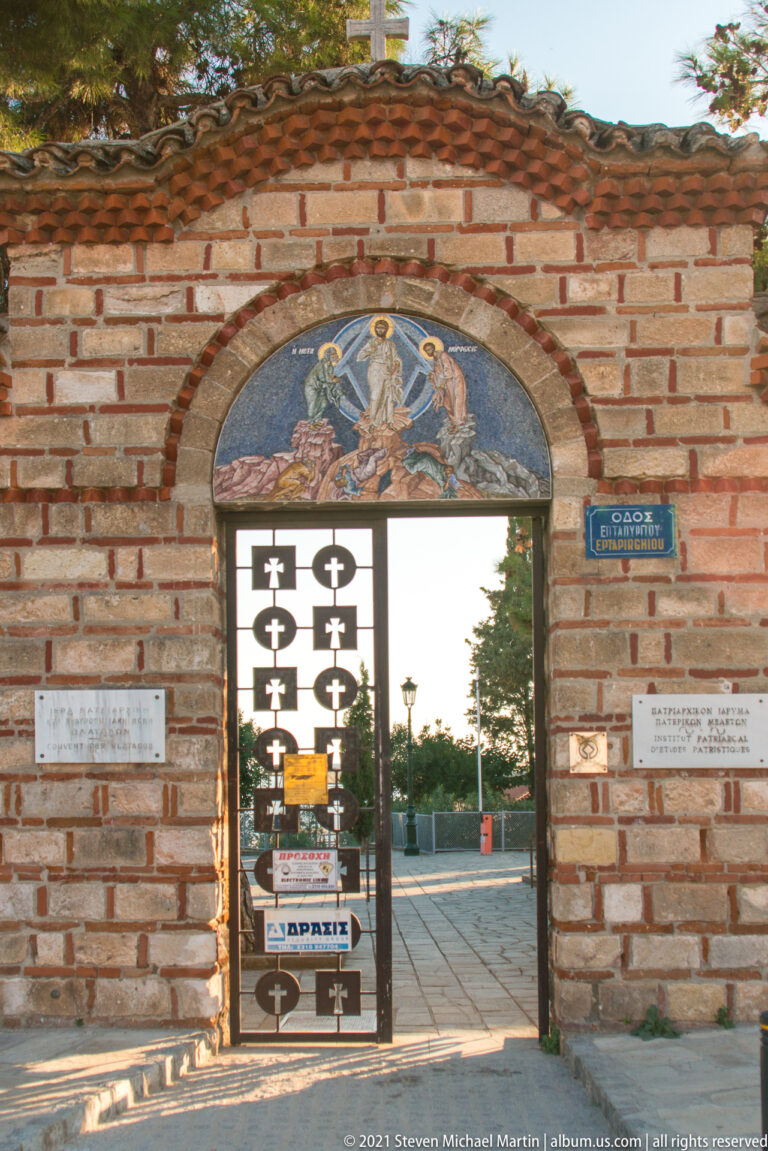 Brick arched gateway with Transfiguration mosaic above a metal gate decorated with cross-shaped cutouts.