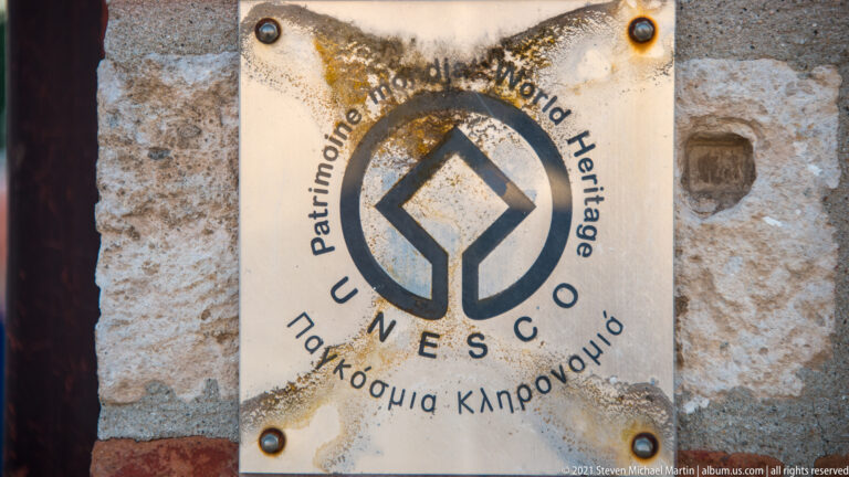 Weathered UNESCO World Heritage plaque with emblem, English wording, and Greek text mounted on stone wall.