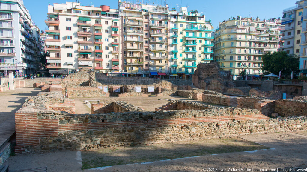 Wide view across low Roman Forum foundations and brick ruins enclosed by modern Thessaloniki apartment blocks.