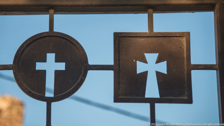 Two cross-shaped cutouts, one in a circle and one in a square, silhouetted in a metal gate against blue sky.