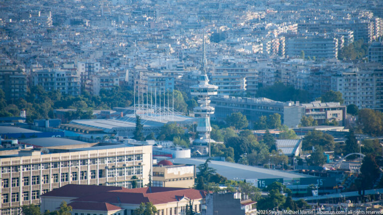 Dense Thessaloniki cityscape with apartment blocks, waterfront buildings, masts, and port infrastructure.