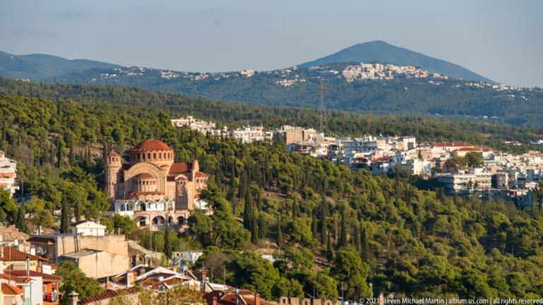 Domed brick church among houses and trees below a broad forested ridge in Thessaloniki.