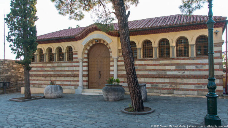 Small striped-masonry church with arched windows, central doorway, courtyard trees, and stone planters.