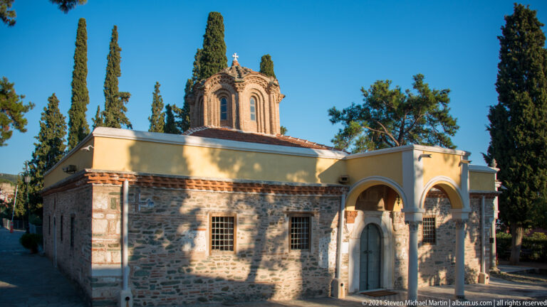Small stone-and-plaster Orthodox church with arched entrances, barred windows, and an open bell turret.