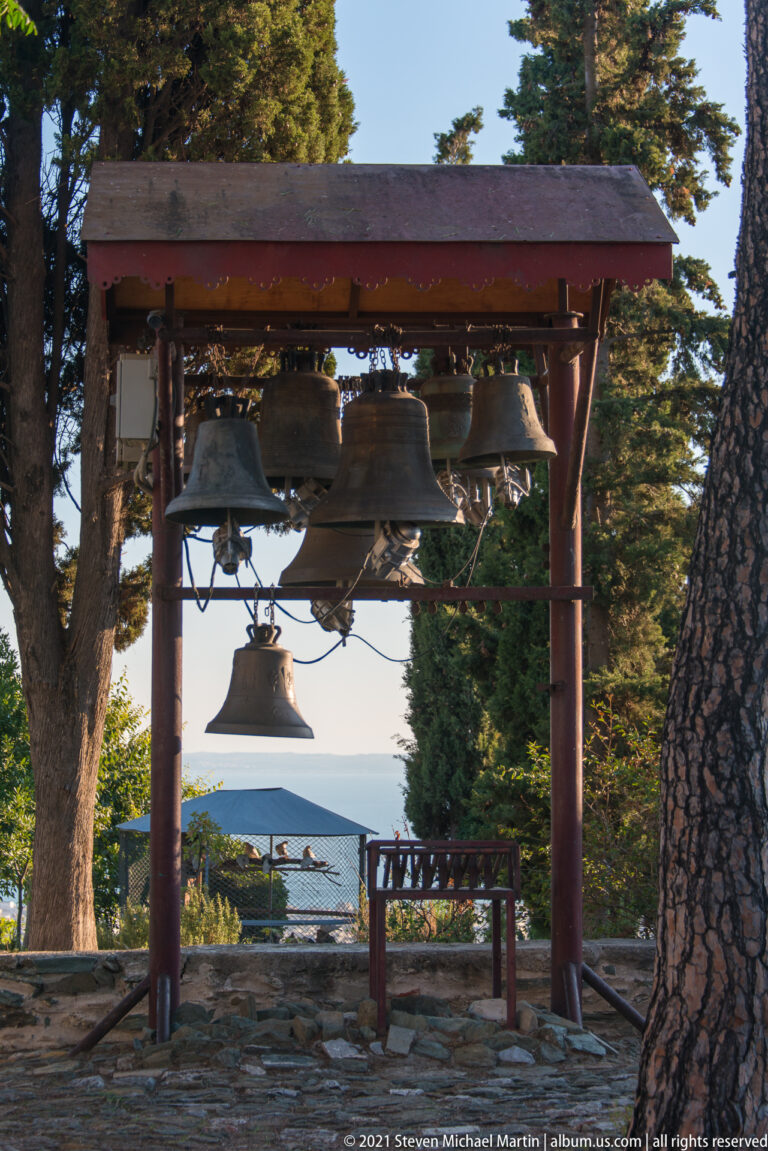 Multiple bronze church bells hanging from an outdoor frame among trees, with a distant city view beyond.