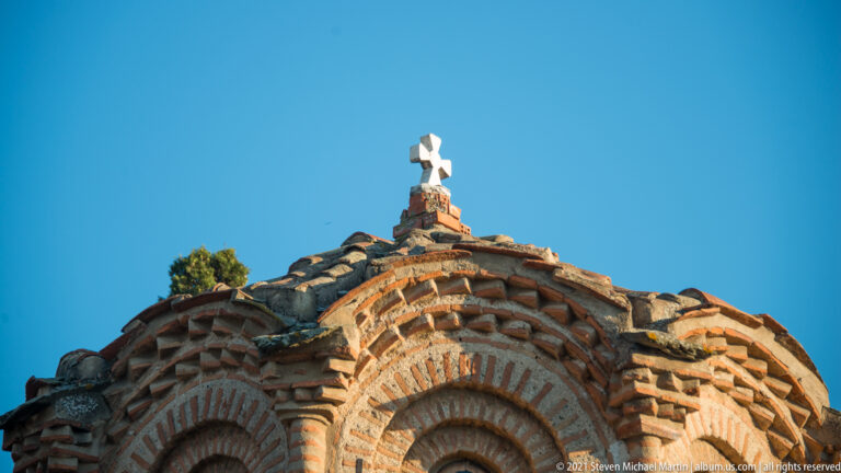 Close view of decorative brick arches and roofline topped by a small white cross against blue sky.