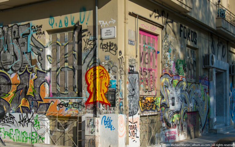 Graffiti, tags, stickers, and painted figures covering shutters and walls on a Thessaloniki street corner.