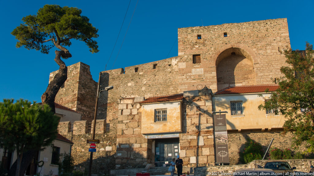 Tall arched gateway in a massive acropolis tower above lower masonry and neighborhood buildings.