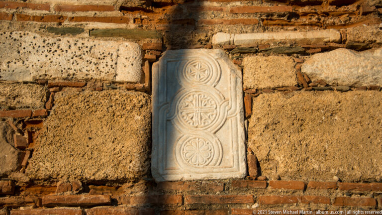 Vertical marble slab with carved circular rosettes reused in rough brick-and-stone wall masonry.