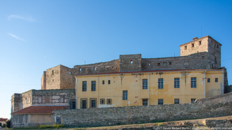 Yellow institutional building enclosed by high stone fortress walls and a square tower at Thessaloniki's upper citadel.