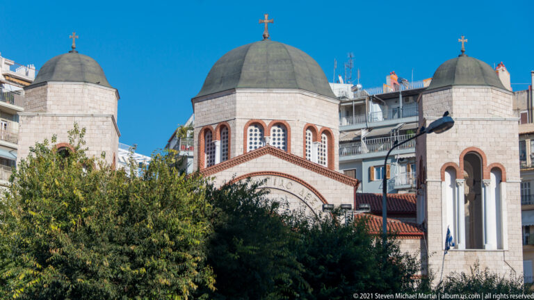 Orthodox church with three dark domes, arched windows, crosses, and pale stone walls above trees.