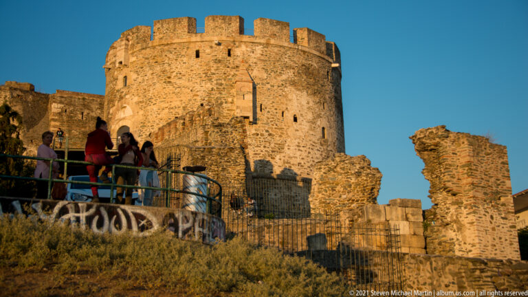 Visitors on a walkway below a large crenellated stone tower and ruined wall.