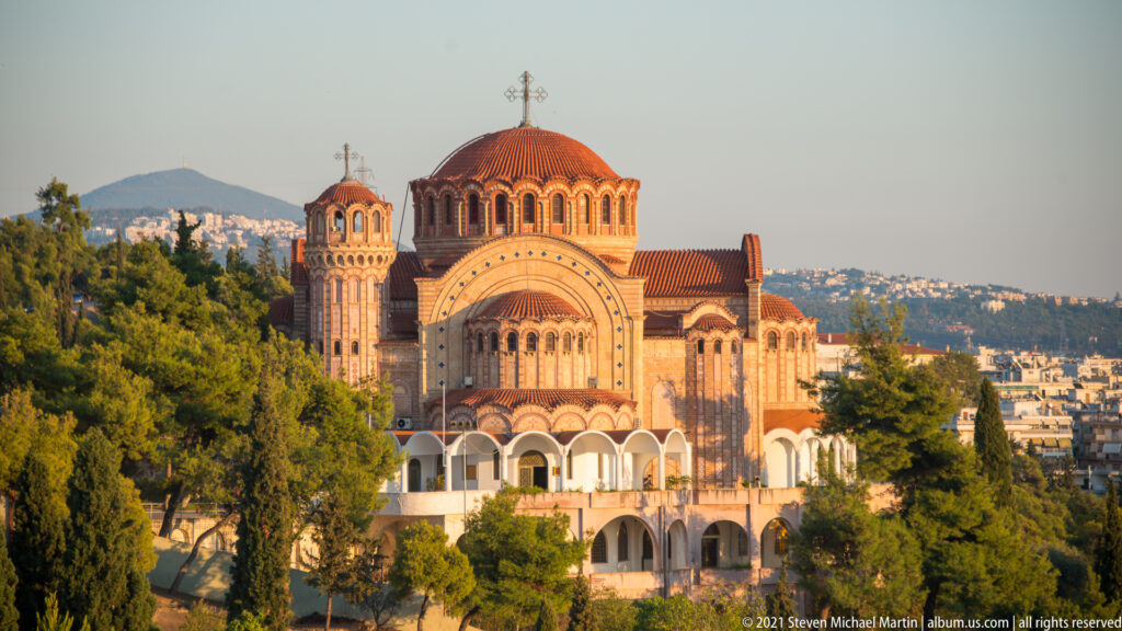 Large domed Orthodox church with bell tower rising above trees and Thessaloniki’s cityscape.